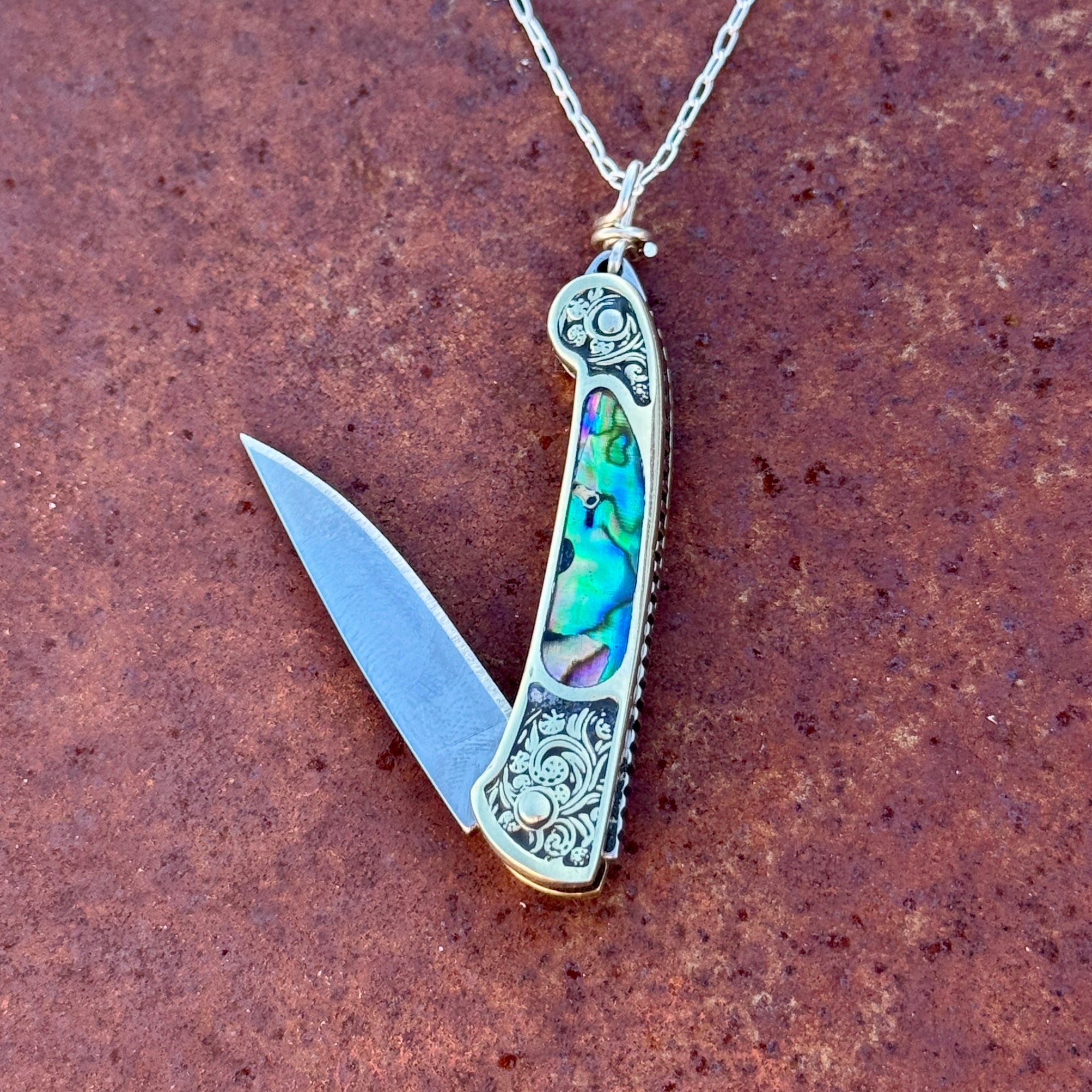 The Lennox Necklace by Fluff Hardware is a functional piece of jewelry featuring a small, ornate pocket knife with gold details and colorful abalone shell inlay—style meets utility in this standout accessory.